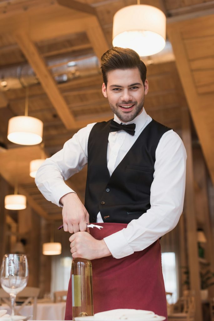 Smiling waiter opening bottle of white wine with corkscrew